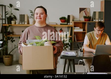 Medium shot of new young female worker with down syndrome standing in office holding her box with personal belongings Stock Photo