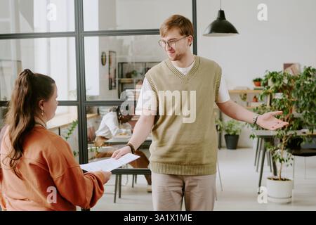 Wide shot of male and female colleague communicating during work in office, unrecognizable woman giving man clipboard with documents Stock Photo