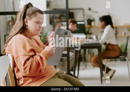 Medium shot of female office worker with down syndrome sitting on chair holding clipboard and filling in documents while her colleagues discussing business plan Stock Photo