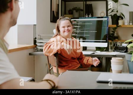 Wide shot of female programmer with down syndrome sitting on chair and looking at her male coworker Stock Photo