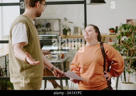Confident female coworker with down syndrome taking documents to her tall male colleague while they standing in center of office Stock Photo
