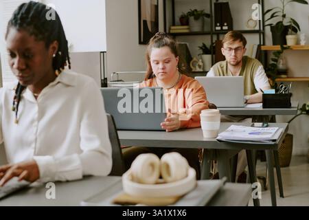 Wide shot of office with biracial team of young programmers sitting at table and coding, their tables equipped with personal belongings Stock Photo
