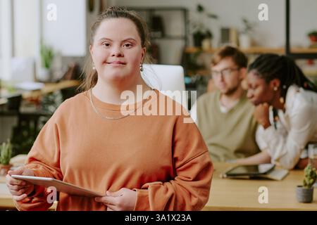 Medium close up portrait of smiling professional female programmer with down syndrome holding white tablet in hands while posing for photo in office Stock Photo