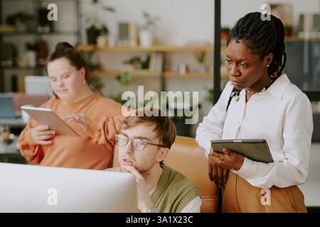 African American female IT specialist with tablet in hands standing near her male colleague while they learning codes on computer monitor Stock Photo