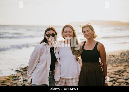 Three women are standing on a beach, smiling and posing for a picture. Scene is cheerful and relaxed, as the women are enjoying their time together at Stock Photo