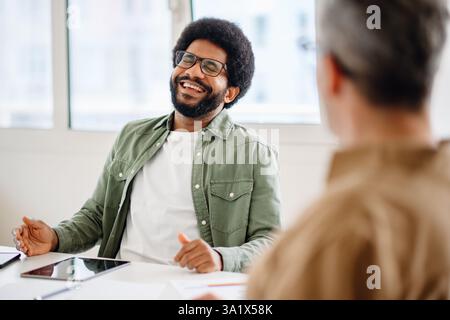 A confident African American businessman in glasses, relaxed and smiling during an office discussion. His body language shows engagement and enthusiasm in a professional environment Stock Photo
