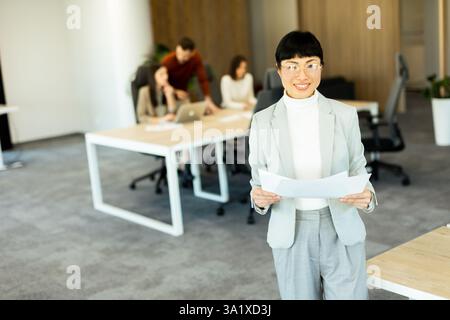 Confident Asian businesswoman stands with documents in hand, smiling at colleagues engaged in discussion at a modern office. Collaboration and creativ Stock Photo