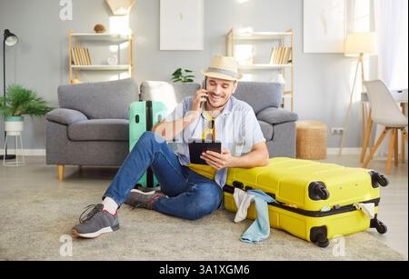 Happy young traveler surrounded by suitcases using tablet, talking on phone and planning trip. Stock Photo
