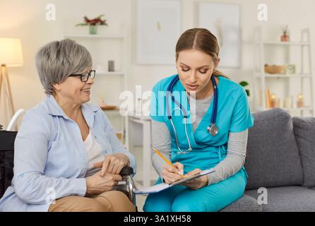 Young nurse taking notes while consulting and listening smiling senior woman in wheelchair at home. Stock Photo