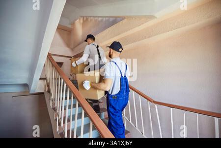 Team Of Movers Carrying Boxes In Stairwell During Relocation To Home Or Office Stock Photo