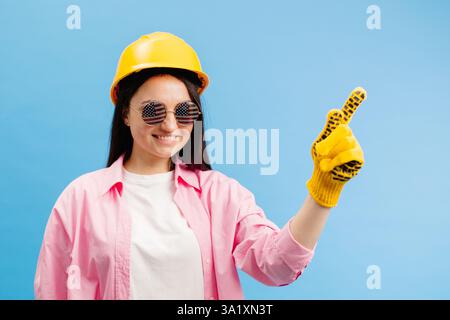 Woman in yellow helmet and gloves pointing and gesturing against blue studio background. Stock Photo