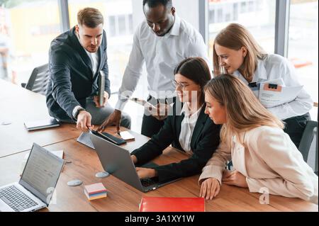 Using laptop that is on the table. Group of office workers are together indoors. Stock Photo
