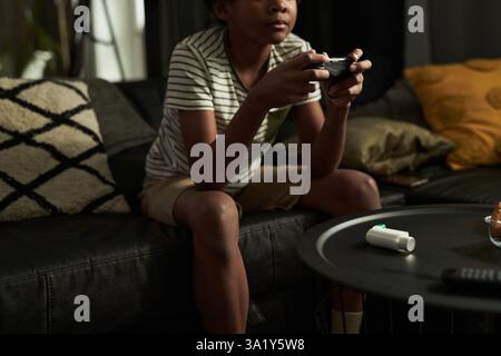 Table with inhaler standing in front of African American asthmatic child with gamepad sitting on sofa and playing video game Stock Photo
