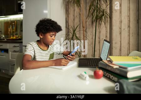 Serious boy with mobile phone looking through online data while sitting in front of laptop and taking notes on distant learning Stock Photo