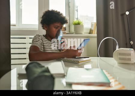 Serious boy with oxygen mask on nose looking at smartphone screen while texting or watching online lesson at home Stock Photo