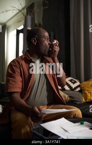 Senior man in casualwear using inhaler during asthma attack while sitting by table with unpaid bills and holding one of them Stock Photo