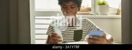 Header of asthmatic schoolboy with inhaler breathing vapors from mask while sitting by table at home and doing schoolwork Stock Photo
