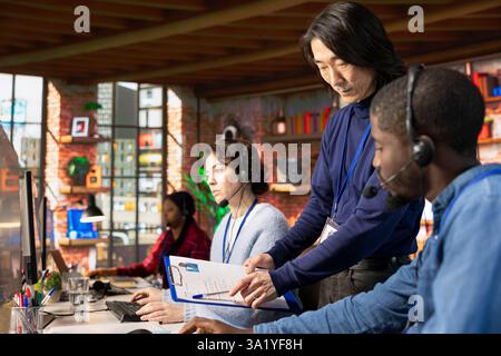 Call center senior manager going over job description file with employee at PC desk. Asian customer support onboarding specialist explaining tasks to intern accommodating with job Stock Photo