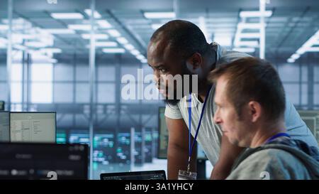 Programmer requesting help from manager while working on computer in data center, monitoring hardware energy consumption. Team leader overseeing IT expert checking for system failures, camera A Stock Photo