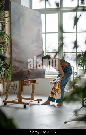 Calm artistic female working on painting in bright creative studio among green plants and flowers. Stock Photo