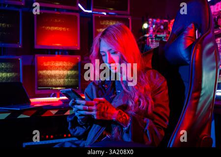Asian woman in front of a computer monitor display using mobile phone, neon light, Hacker wear black Stock Photo