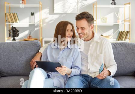 Young couple sitting on sofa, using digital tablet at home, enjoying entertainment and video calls. Stock Photo