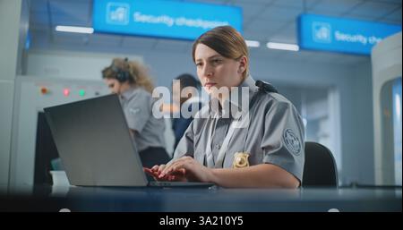 Airport Security: Female TSA Officer Using Laptop, Smiling and Looking at Camera. Transportation Security Administration for Travel Safety. In Background, Airport Staff Conducting Screening Procedures Stock Photo