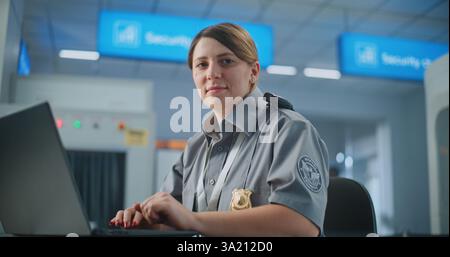 Airport Security: Female TSA Officer Using Laptop, Smiling and Looking at Camera. Transportation Security Administration for Travel Safety. In Background, Airport Staff Conducting Screening Procedures Stock Photo