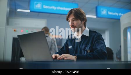 Airport Security Check: Portrait of TSA Worker Using Laptop, Looking at Camera. Transportation Security Administration for Travel Safety. In Background, Airport Staff Conducting Screening Procedures. Stock Photo