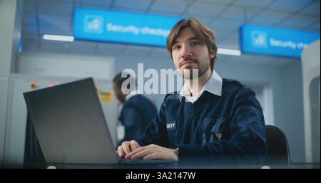 Airport Security Check: Portrait of TSA Worker Using Laptop, Looking at Camera. Transportation Security Administration for Travel Safety. In Background, Airport Staff Conducting Screening Procedures. Stock Photo