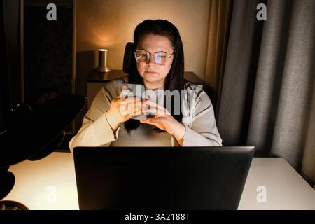 Serious woman freelancer with eyeglasses using smartphone while working with laptop Stock Photo