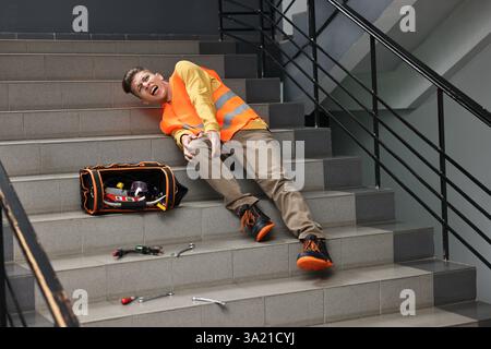 Accident at work. Injured man with toolkit on stairs indoors Stock Photo