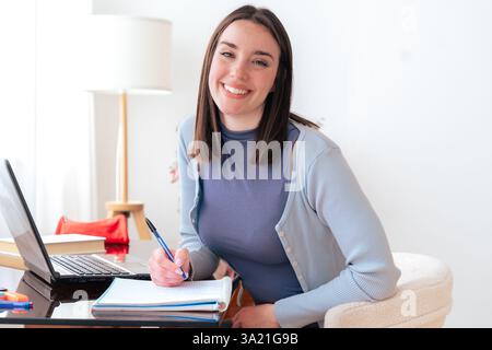 Smiling caucasian young woman working on laptop at home office with notebooks Stock Photo