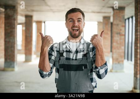 With thumbs up, smiling. Construction worker in uniform in empty unfinished room. Stock Photo
