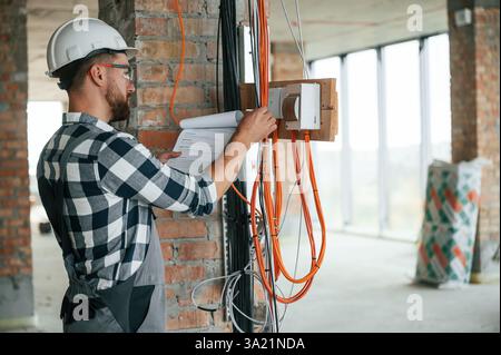 Working with electricity. Construction man in uniform in empty unfinished room. Stock Photo