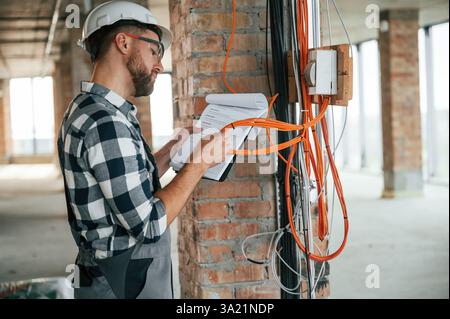 Working with electricity. Construction man in uniform in empty unfinished room. Stock Photo