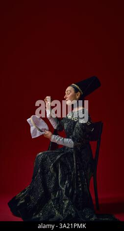 Elderly woman in historical noble dress carefully embroidering fabric with concentrated expression, hands gracefully working against deep red Stock Photo