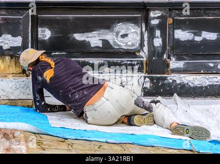 Young woman decorator lying on dustsheet on pavement whilst renovating shop front woodwork - Loches, Indre-et-Loire (37), France. Stock Photo