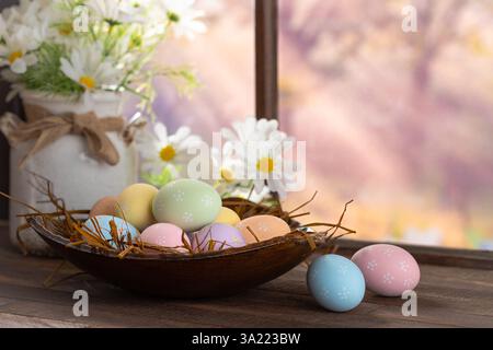 Easter eggs in a bowl with white daisies on a wooden table next to window with colorful springtime background Stock Photo