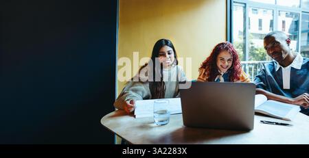 Three multicultural students studying together on campus, working with a laptop and textbooks, showcasing teamwork and collaboration. Stock Photo