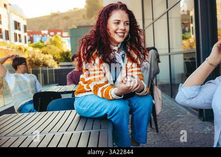 A cheerful female student with curly hair sitting on a table at a city college campus, enjoying a sunny day with friends and vibrant surroundings. Stock Photo