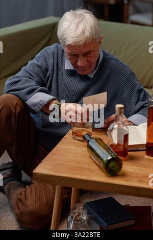 Elderly man sitting on sofa intently reading paper while holding glass of whiskey, with bottle and papers on table, creating a contemplative atmosphere Stock Photo