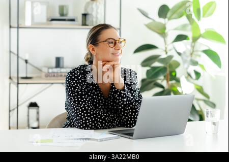 Positive pensive businesswoman with glasses, company manager, secretary, sitting at a desk with a laptop in the office, thoughtfully looking away, smiling and dreaming about something good Stock Photo