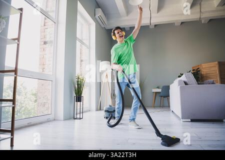Young man having fun while vacuuming in a modern daylight-filled living room, showcasing casual style and a joyful mood. Stock Photo