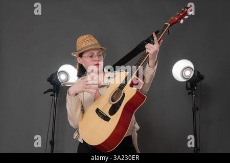 Playful woman strummed classical acoustic guitar, creating an exuberant, lively atmosphere. Musician wearing eyeglasses, leather jacket and stylish st Stock Photo