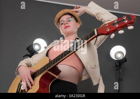 Self assured female guitarist is absolutely thrilling to look at. She's holding acoustic guitar, looking at camera. One hand cradles brim of straw hat Stock Photo