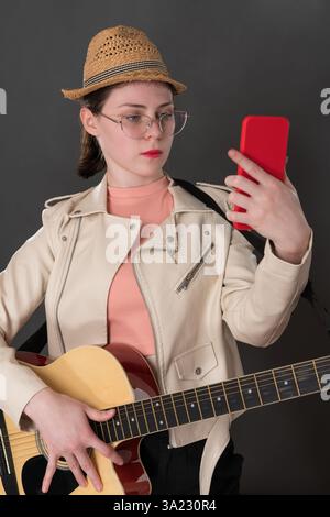 Young woman guitarist with guitar using cell phone, looking at portable information device while learning to play six string acoustic guitar. Modern t Stock Photo