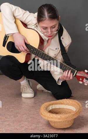 Female street musician strumming six string acoustic guitar for money and in front of her lying empty straw hat for donation for playing guitar. Woman Stock Photo