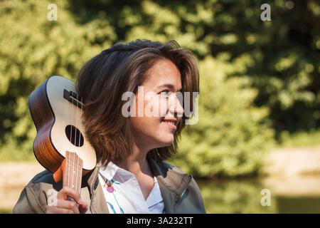 Smiling woman enjoying peaceful moments in nature while practicing ukulele. Happy young female musician outdoors Stock Photo