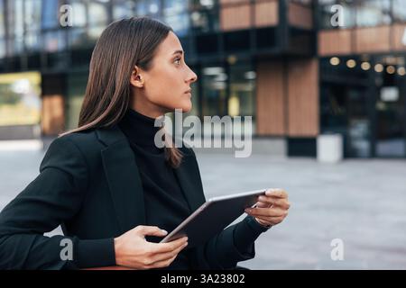 Side view portrait of young businesswoman with black hair sitting outside looking away. Female with digital tablet sitting outside against office buil Stock Photo